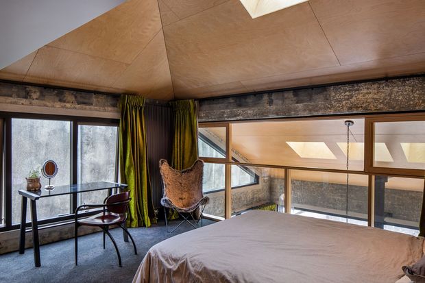 A distinctive bedroom with a low, warm timber-panelled ceiling and exposed weathered structural beams. The room features a large bed, dark carpet, full-height glazing and internal windows overlooking a lower-level space, with a compact glass-topped desk, sculptural chair, and deep green curtains. Skylights and clerestory-style glazing bring in natural light, creating a dramatic, loft-like interior.