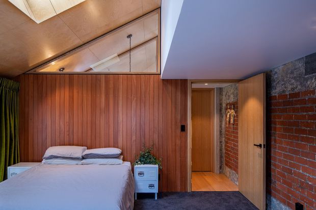 A warm, compact bedroom with a bed positioned against a full-height vertical timber-panelled feature wall. The room includes a small white bedside cabinet, dark carpet, green curtains, and a timber-lined ceiling with a mirrored or reflective inset. An open timber door leads to a hallway, while exposed red brick and rough stone wall surfaces add an industrial character.