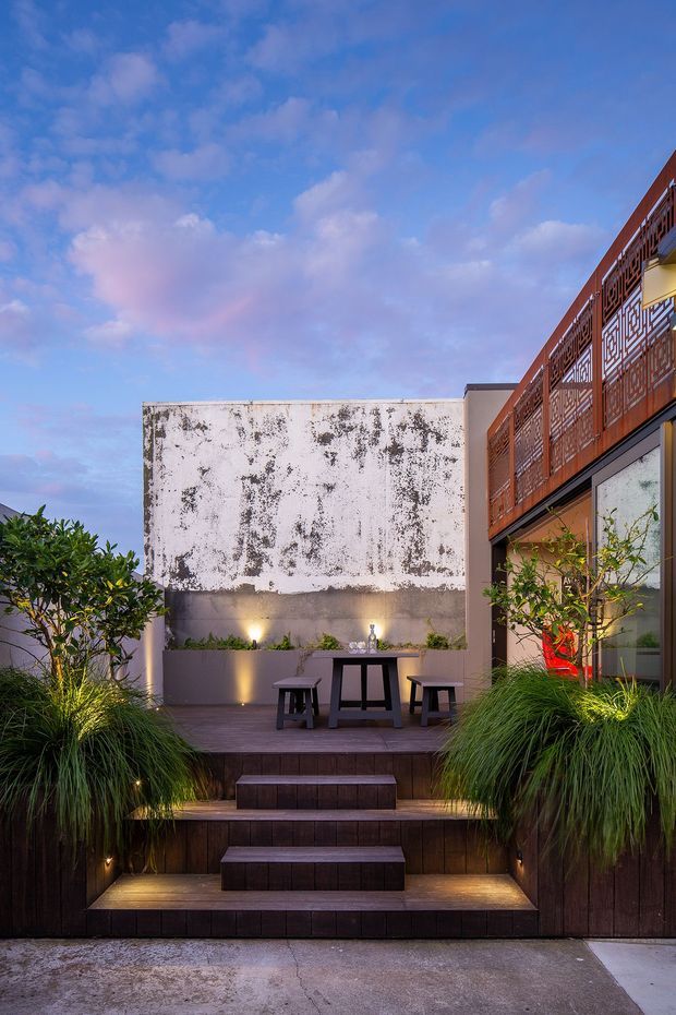 A landscaped urban courtyard and raised timber deck viewed from outside, with broad illuminated steps leading to a compact outdoor dining area. The space includes a dark timber-clad deck and retaining walls, built-in planter beds with ornamental grasses and small trees, a simple outdoor table with stools, wall uplighting, and a weathered rendered wall backdrop. A multi-storey building with large glazing and distinctive reddish-brown geometric metal balcony screening borders the right side. The image is photographed at dusk under a blue and pink sky.