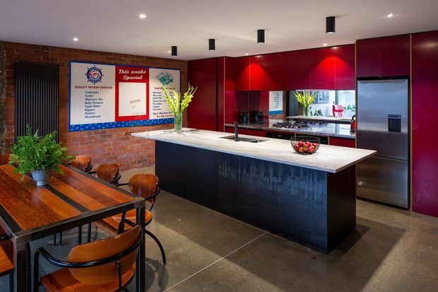 A contemporary open-plan kitchen and dining area with deep red glossy cabinetry, a large dark island with a pale stone benchtop, stainless-steel appliances, and a polished concrete-look floor. Exposed red brick walls add an industrial character, while a timber dining table and curved timber chairs occupy the foreground. Black recessed and pendant-style ceiling lights, a mirrored splashback, and indoor greenery complete the space.
