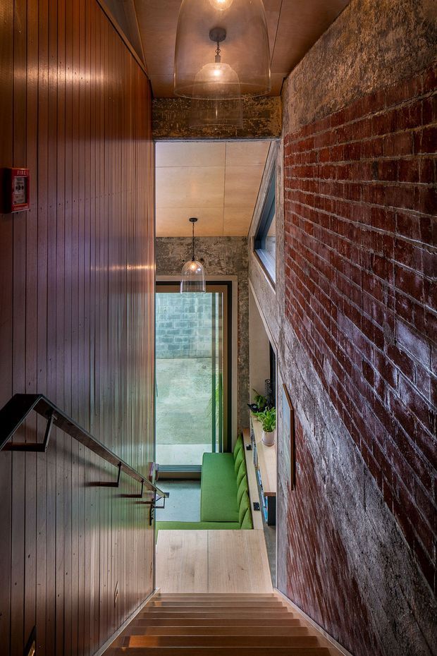 A narrow interior stairway descending alongside an exposed red-brick wall and dark vertical timber-panelled wall. Warm pendant lights hang from a timber-lined ceiling, while a green upholstered built-in bench and a tall glazed door or window bring natural light into the lower landing. The space has an industrial, adaptive-reuse character with strong warm timber and masonry textures.