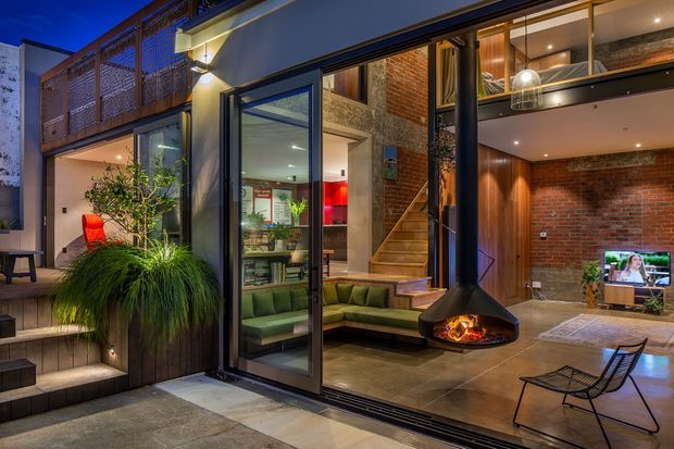 A dramatic dusk view into a contemporary multi-level living room through fully opened glazed doors. The interior features exposed red brick walls, warm timber wall lining, polished concrete flooring, a suspended black fireplace with an active fire, built-in green seating, a television, and a staircase leading to an upper level. The adjoining exterior terrace has stepped paving, lush ornamental planting, and a balcony with patterned screening. The composition emphasises indoor-outdoor connection and double-height volume.