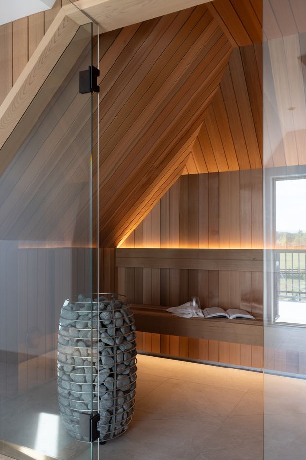 A warmly lit sauna interior enclosed by clear glass, featuring extensive diagonal timber wall and ceiling panelling, an integrated timber bench, a cylindrical stone-filled sauna heater, and a pale tiled floor. A window provides daylight at the right.