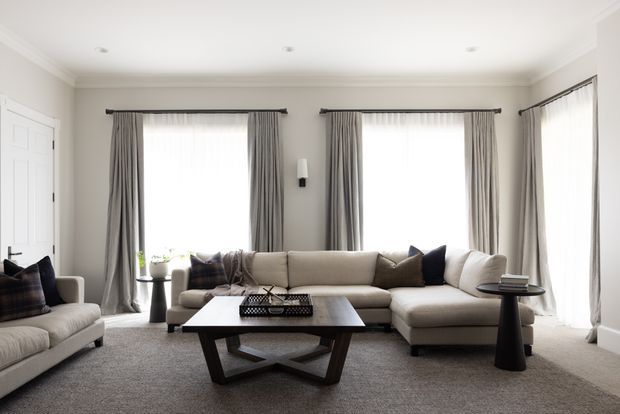 A bright, spacious formal living room with a large L-shaped upholstered sectional, matching armchair, dark geometric coffee table, and side table. The room has pale neutral walls, full-height grey curtains across multiple windows, recessed ceiling lights, decorative crown moulding, and soft grey carpeting. The overall look is refined and contemporary with traditional architectural detailing.