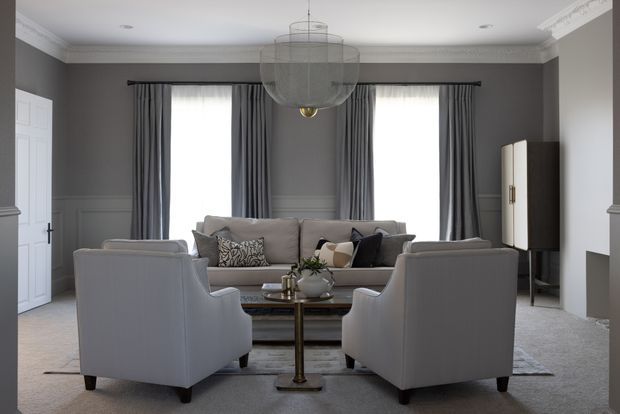 Elegant formal living room with grey paneled walls, ornate white crown moulding, full-height dark grey curtains, plush grey seating arranged around a central coffee table, and a large tiered glass pendant light. A dark timber cabinet and fireplace add contrast to the refined, symmetrical space.