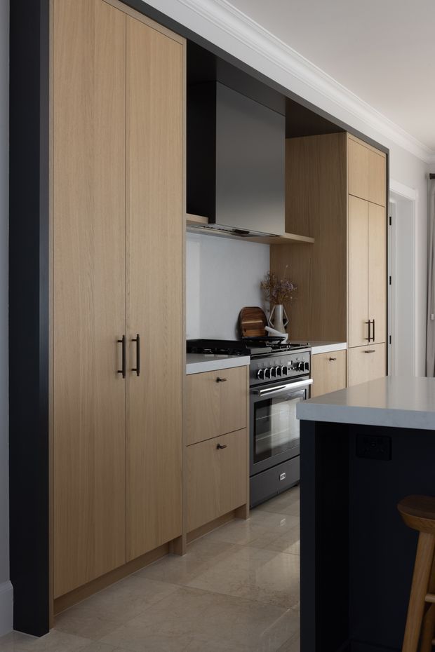 A refined contemporary kitchen with full-height light timber cabinetry, integrated storage, a stainless-steel range cooker and hood, pale stone benchtops, and a dark island with seating. The cabinetry is framed by a dark portal, while white decorative ceiling cornices and a light tiled floor add contrast.