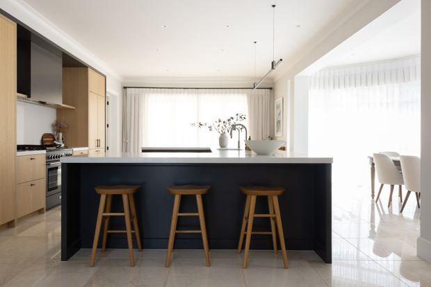 Bright contemporary open-plan kitchen with a large dark island and three timber stools, light timber cabinetry, integrated stainless-steel appliances, pale stone-look flooring, and a dining area visible to the right. Floor-to-ceiling sheer curtains and abundant natural light create an airy, refined interior.