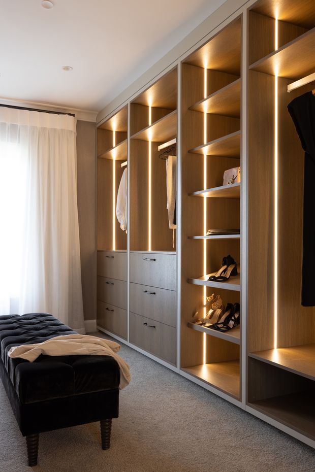 A refined bedroom dressing area with a full wall of custom built-in wardrobes and open shelving. The joinery is finished in warm wood veneer with integrated vertical LED lighting, drawers, hanging rails, and shoe storage. A dark upholstered bench sits on a soft grey carpet beside sheer curtains and a bright window.
