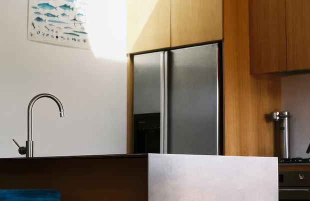 A compact contemporary kitchen viewed across a dark island or peninsula, with a tall curved stainless-steel tap, warm timber cabinetry, integrated dark-framed refrigerator, and dark benchtops. A partial dining chair and fish artwork are visible, with strong sunlight creating a warm, relaxed atmosphere.
