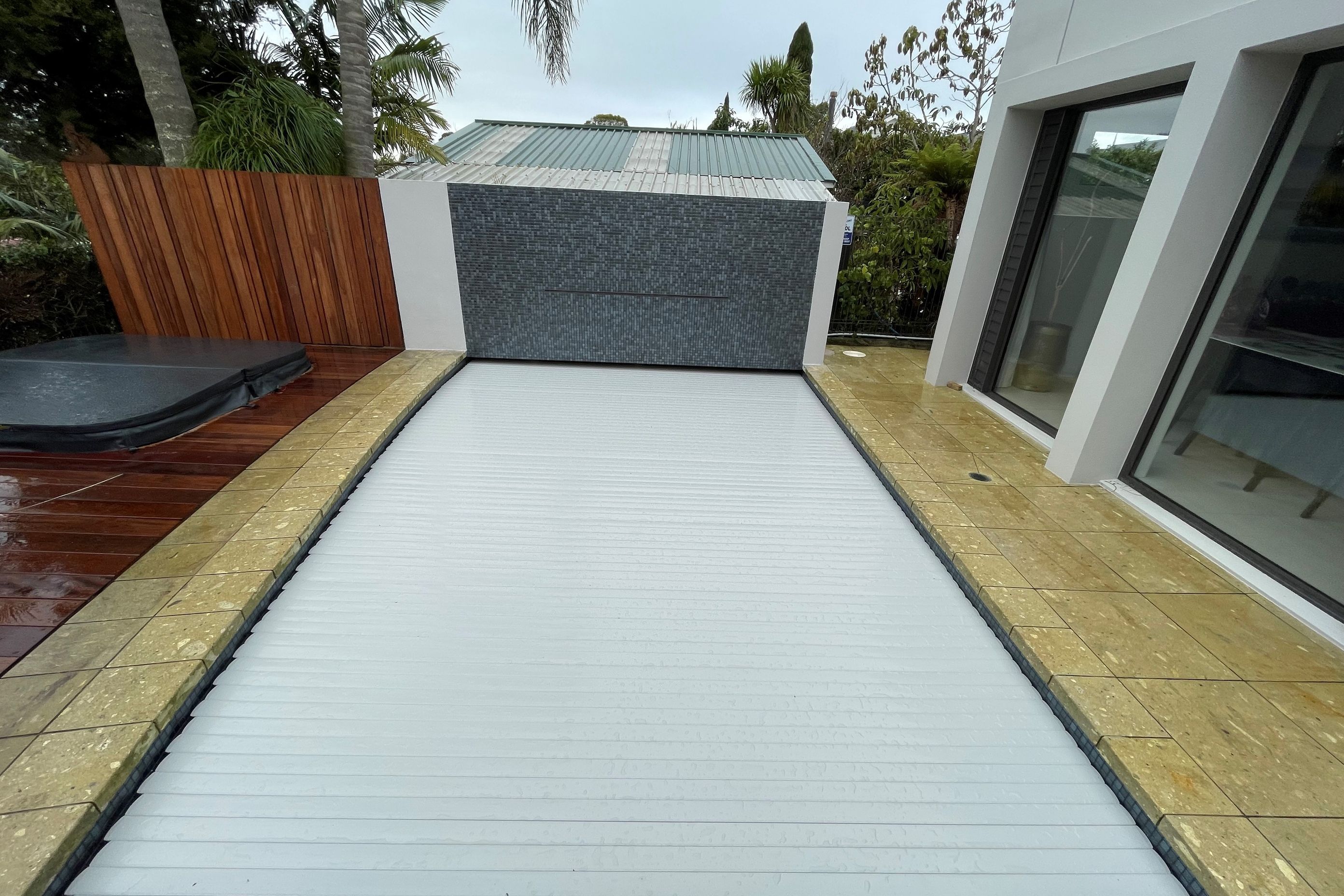 After Renovation - The Rear Feature wall was  'squared up' and fully tiled.  The addition of a rain arch water feature adds interest and ambience.  The existing slatted pool cover was retro fitted with new slats from BMH.  The Pool was fully tiled with Ezarri Capricon 25mm Tiles - looks very striking.