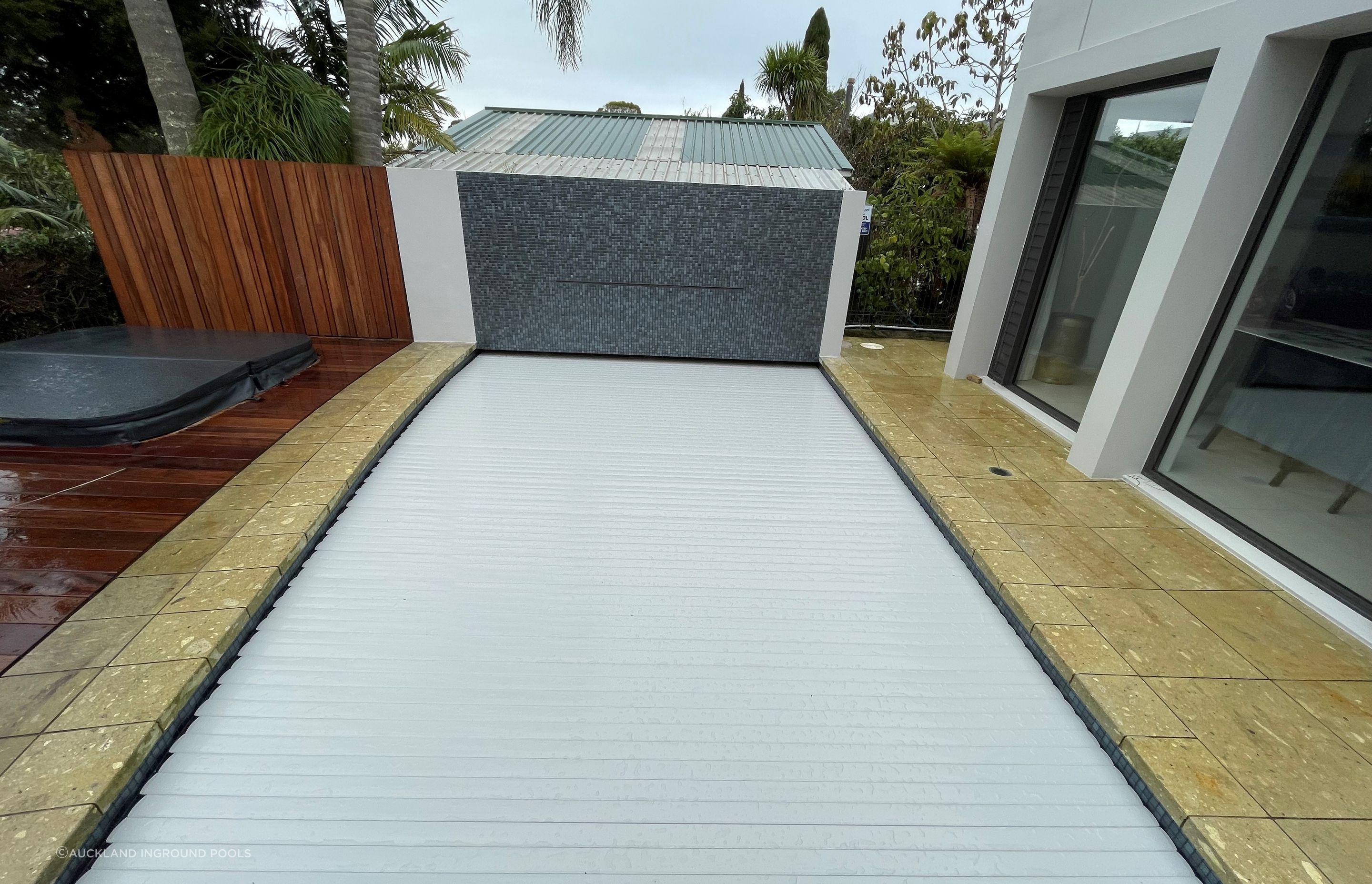 After Renovation - The Rear Feature wall was  'squared up' and fully tiled.  The addition of a rain arch water feature adds interest and ambience.  The existing slatted pool cover was retro fitted with new slats from BMH.  The Pool was fully tiled with Ezarri Capricon 25mm Tiles - looks very striking.