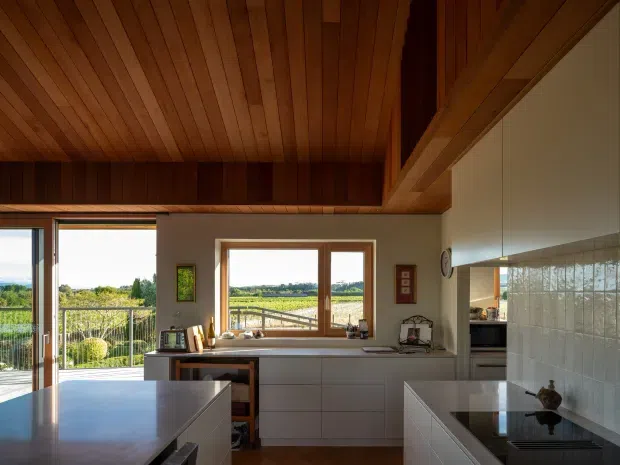 Warm, light-filled contemporary kitchen with extensive white cabinetry, two dark stone-topped islands or counters, a tiled splashback, and a dramatic timber-lined sloping ceiling with exposed structural beams. Large windows and an open glazed door provide views of a green rural landscape and outdoor deck or balcony.