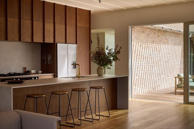 Warm, open-plan kitchen with a long island and integrated breakfast bar, featuring dark timber vertical cabinetry, a pale stone benchtop, stainless-steel appliances, and timber flooring. The kitchen opens to a bright outdoor deck or courtyard through large glazed doors, with a glimpse of a furnished living area beyond.