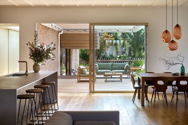 Warm, open-plan residential interior with a contemporary kitchen, dining area and living space opening onto a timber deck through large glazed sliding doors. The kitchen features a long pale stone island with dark stools and brass-toned tapware, while the dining area has a timber table, sculptural pendant lights and built-in-looking timber joinery. A green sofa sits outside on the deck, with lush planting visible beyond.