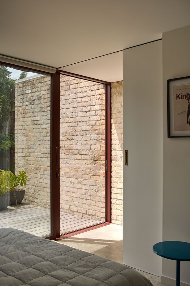 A sunlit bedroom interior with a low-profile bed in the foreground, opening through large glazed doors onto a timber deck and enclosed brick courtyard. The glazing has dark reddish-brown timber frames, with a white wall, framed artwork, and a small round blue side table visible inside. The space has a simple modern composition and strong indoor-outdoor connection.