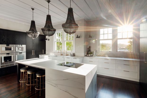 A bright, high-end kitchen with a large white marble-look island, integrated cooktop, and three bar stools. The room features dark timber cabinetry and flooring, white shaker-style perimeter cabinetry, a white planked ceiling, large multi-pane windows, stainless-steel appliances, and oversized decorative pendant lights.
