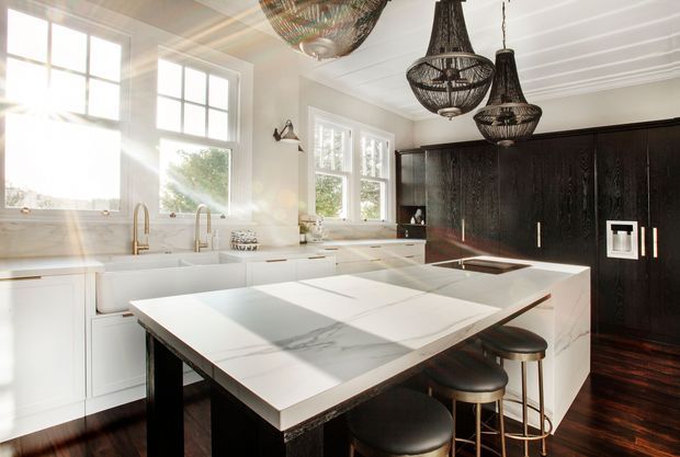Bright, spacious kitchen with a large white marble-look island, integrated seating, white cabinetry, farmhouse sink, brass-toned fixtures, and a contrasting dark timber wall of cabinetry. Decorative woven pendant lights and a coffered or panelled white ceiling add character, while large multi-pane windows provide abundant natural light.