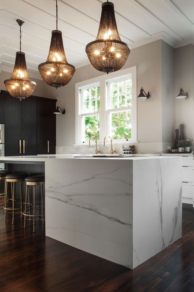 A refined kitchen featuring a large waterfall-edge marble or engineered-stone island, dark built-in cabinetry, white cabinetry, brass-toned fixtures, dark timber flooring, and three oversized woven pendant lights. White-framed windows and pale walls create a bright yet warm, classic interior.