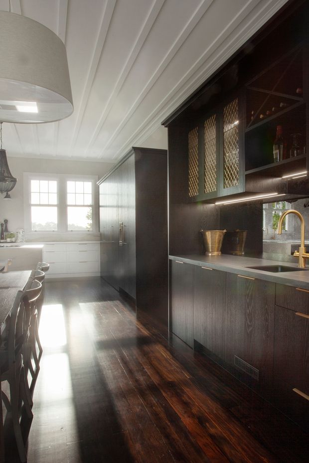 A long, refined kitchen with dark timber cabinetry, a dark stone benchtop and integrated sink, brass tapware, illuminated glass-front upper cupboards, and a white painted timber-lined ceiling. A dining area with stools sits to the left, while bright multi-pane windows and white cabinetry bring natural light into the space. Dark polished timber flooring runs throughout.