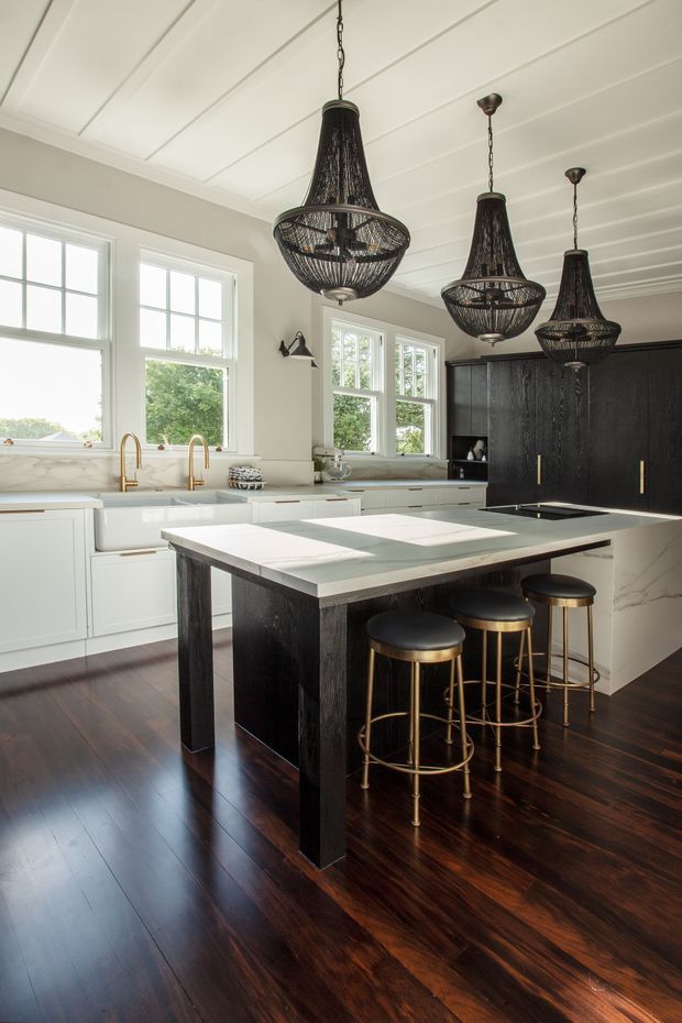 A bright, refined kitchen with a large central island, white cabinetry, black textured joinery, dark timber flooring, marble-look benchtops, brass tapware and stools, and three oversized woven pendant lights. Large multi-pane windows and a white panelled ceiling bring in abundant natural light.