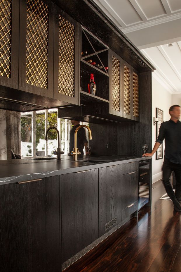 A sophisticated dark-toned kitchen and home bar area with black-stained timber cabinetry, brass diamond-grille door inserts, a dark stone benchtop, twin curved brass and black faucets, mirrored splashback, open bottle shelving, and polished dark timber flooring. White coffered ceiling detailing and a bright window provide contrast.