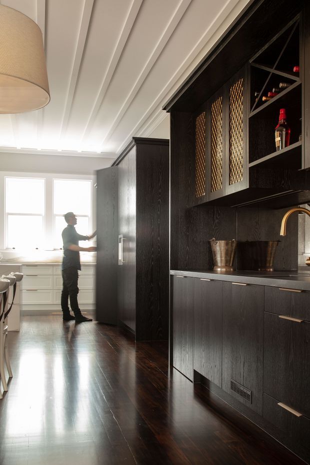 A refined galley kitchen with dark timber cabinetry and matching wall panelling, polished dark timber flooring, a pale stone or composite benchtop, and a brass gooseneck tap. Open shelving and decorative metal-grille cabinet fronts create a bar or drinks-station feel, while a bright secondary kitchen area with white cabinetry is visible at the far end. The ceiling has distinctive white linear panelling and integrated lighting.