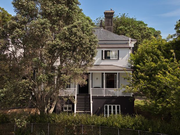 Front exterior view of a multi-storey heritage-style house surrounded by mature trees and garden planting. The facade features painted horizontal weatherboards, a steep hipped roof, prominent masonry chimney, and a covered upper-level veranda with white balustrading. Dark lower-level cladding and steps contrast with the light upper facade.