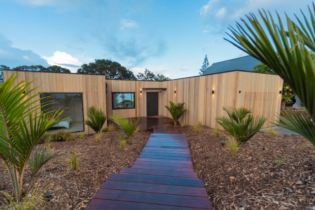 Single-storey contemporary timber-clad building with a low-pitched roof, dark entry door and window frames, and a straight timber walkway leading through a landscaped front garden. The façade is composed of warm vertical timber boards, with palms and drought-tolerant planting surrounding the entrance.