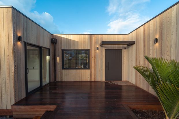 A modern timber-clad courtyard or entry terrace enclosed by low, angular walls. Dark stained timber decking contrasts with vertical light timber wall cladding, black-framed glazing, a dark entry door, and minimalist exterior wall lighting. A palm plant adds a small tropical accent beneath a bright open sky.