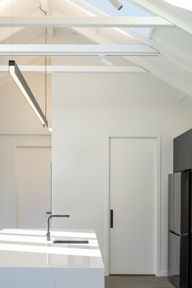 Bright contemporary kitchen interior with a white stone island and integrated sink, black tapware, dark cabinetry and stainless-steel refrigerator. The room has a high vaulted ceiling with exposed white-painted beams, roof skylights, and linear pendant lighting. A concealed door and pantry-style cabinetry are visible at the rear.