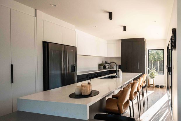 Bright contemporary open-plan kitchen with a large pale stone island and integrated breakfast bar, lined with tan upholstered stools. Full-height white cabinetry, dark appliances and dark timber-toned joinery create contrast, while large glazing at the rear brings in abundant natural light.