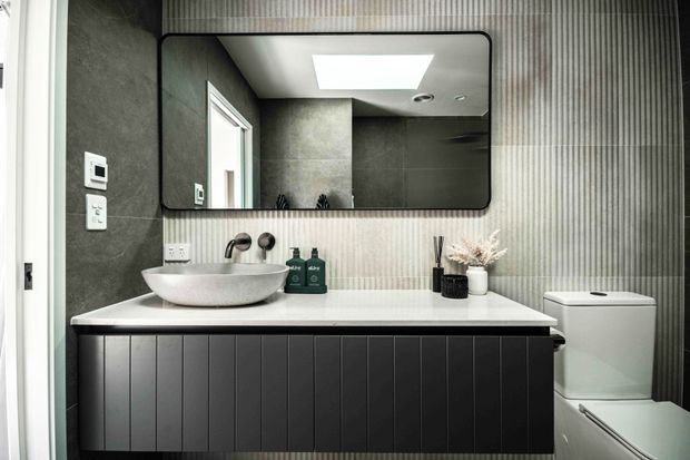 Contemporary bathroom vanity with a white vessel basin, wall-mounted brushed-metal tapware, dark vertical-panel cabinetry, light stone benchtop, and vertically ribbed wall tiling. A large rounded-rectangle mirror reflects the shower area and a bright skylight. The toilet is positioned beside the vanity.