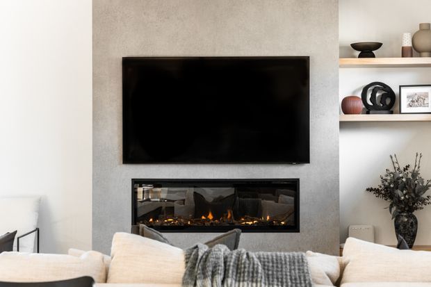 Contemporary living room featuring a wall-mounted flat-screen television above a built-in linear fireplace, set within a smooth light-grey feature wall. Open white display shelving with decorative objects and a vase occupies the right side, while a large pale sectional sofa with a textured throw fills the foreground.