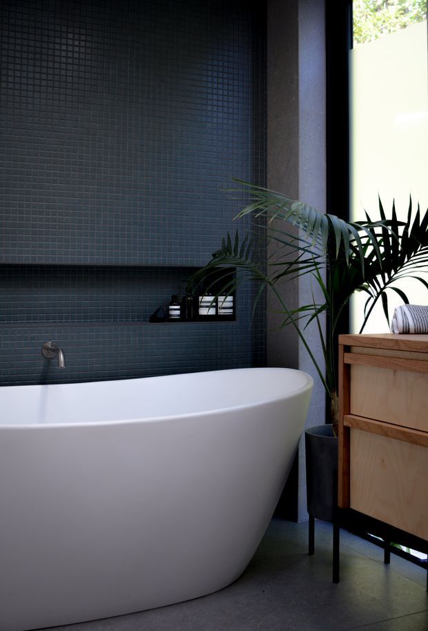 A dark, refined contemporary bathroom featuring a freestanding white bathtub, charcoal-blue small-format tiled walls, a recessed shelf with bath products, a wall-mounted tap, a timber vanity, and lush indoor plants beside a bright window.
