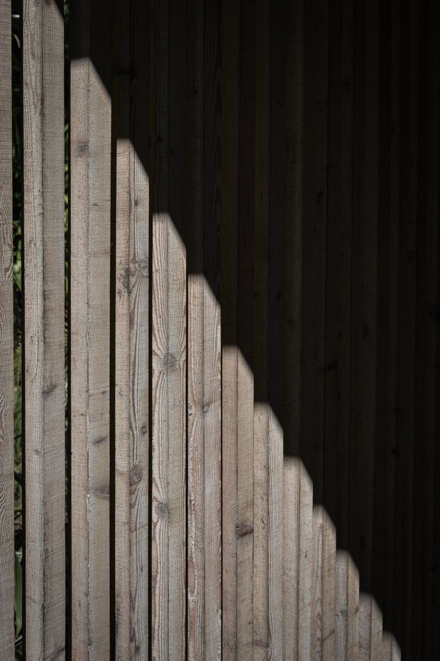 Close-up architectural detail of vertically arranged rough-sawn timber boards, with strong diagonal sunlight and shadow creating a high-contrast geometric pattern across the wood surface. The image is abstract and texture-focused, with no visible furniture or room context.