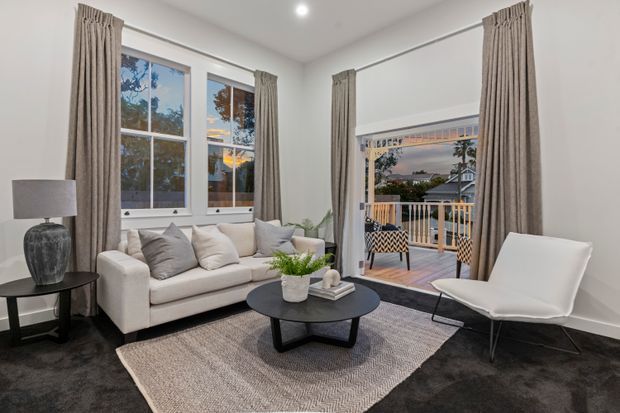 Bright, compact living room with white walls, tall corner windows and a glazed balcony door framed by full-length taupe curtains. The room contains a white sofa with neutral cushions, a sculptural white lounge chair, a black round coffee table, side table and lamp, and a large textured area rug over dark carpet. A balcony and neighbouring residential views are visible beyond the glazing.