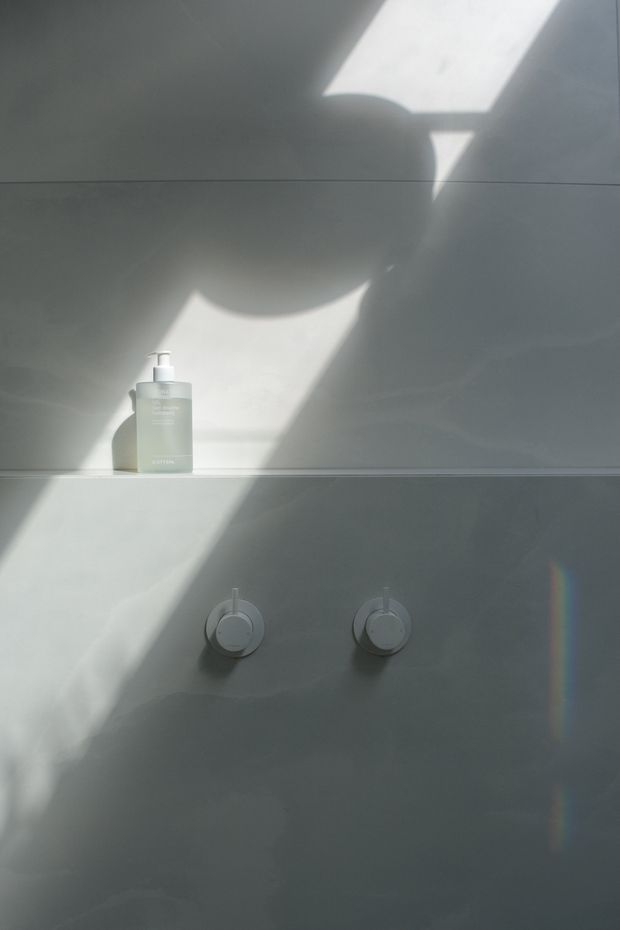 Minimal close-up of a pale tiled bathroom wall with two round wall-mounted shower controls and a small pump soap dispenser on a narrow ledge. Strong diagonal sunlight and shadow patterns create a quiet, atmospheric composition; no broader room layout is visible.