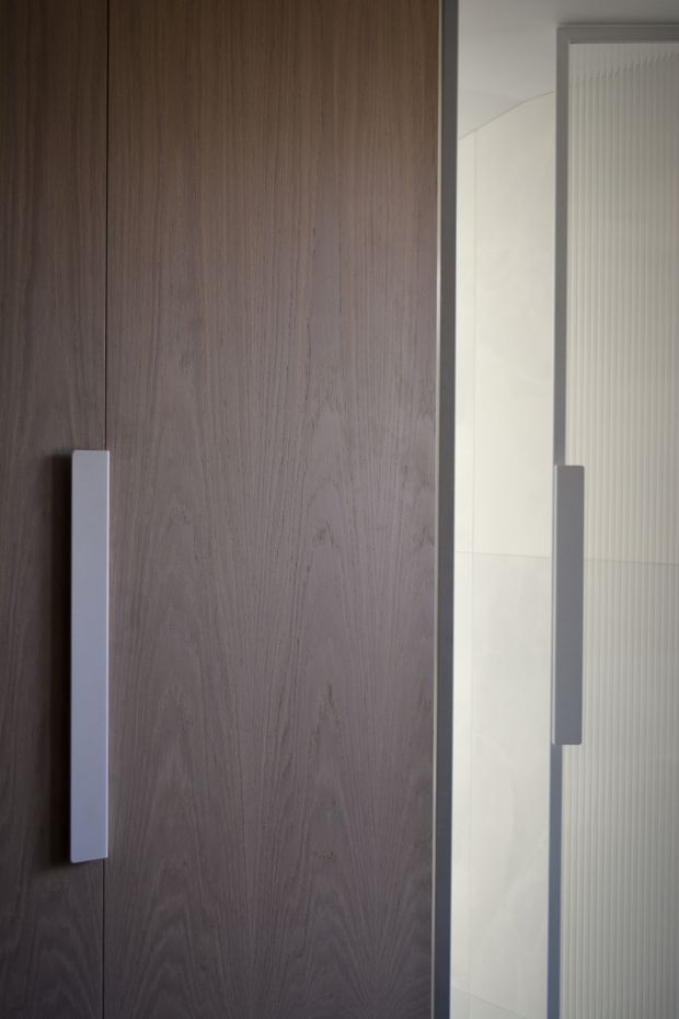 Close-up view of tall fixed joinery with dark stained wood-grain doors and long rectangular light-coloured handles. A partially visible adjacent glazed or ribbed-panel door has a matching vertical handle, creating a clean, understated built-in storage or wardrobe composition.