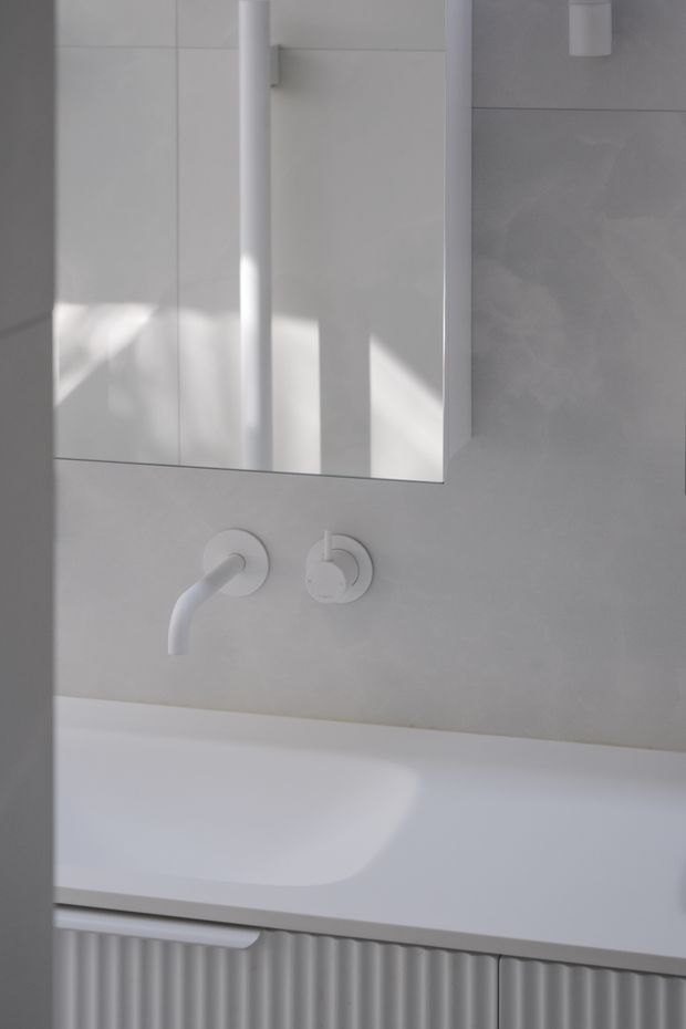 Close-up of a minimalist white bathroom vanity and bathtub area. A wall-mounted white basin faucet and separate circular control sit beneath a rectangular mirrored cabinet. Pale large-format wall tiles, a white integrated benchtop or basin surface, and fluted light-grey cabinetry create a monochromatic, sculptural interior. Strong sunlight casts geometric shadows across the surfaces.