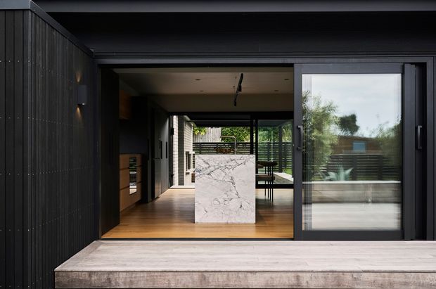 A dark, contemporary residential facade opens onto a covered outdoor deck through large black-framed sliding glass doors. The view looks into an open-plan kitchen and dining area, with a marble-clad island, warm timber flooring, black cabinetry, and a timber privacy screen beyond. A broad concrete or stone terrace step occupies the foreground.