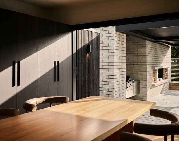 A covered outdoor dining area with a large timber dining table and curved upholstered chairs, adjoining a dark vertical-panel storage wall and a pale brick outdoor fireplace or barbecue structure. Strong sunlight creates angular shadows across the surfaces, with a slatted screen and courtyard glimpsed beyond.