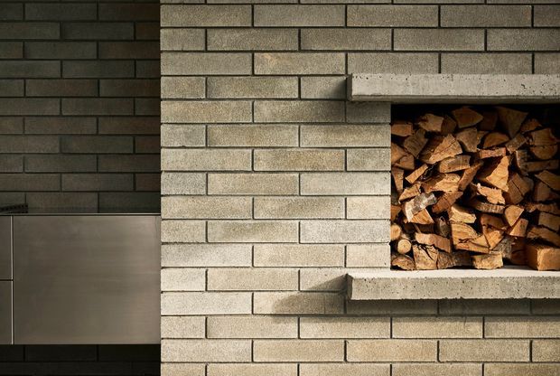 Close-up architectural detail of a contemporary masonry fireplace with a recessed firewood storage niche. Light grey brickwork forms the main wall and concrete shelf edges frame the stacked timber, while a darker charcoal brick wall and smooth metal panel appear to the left.