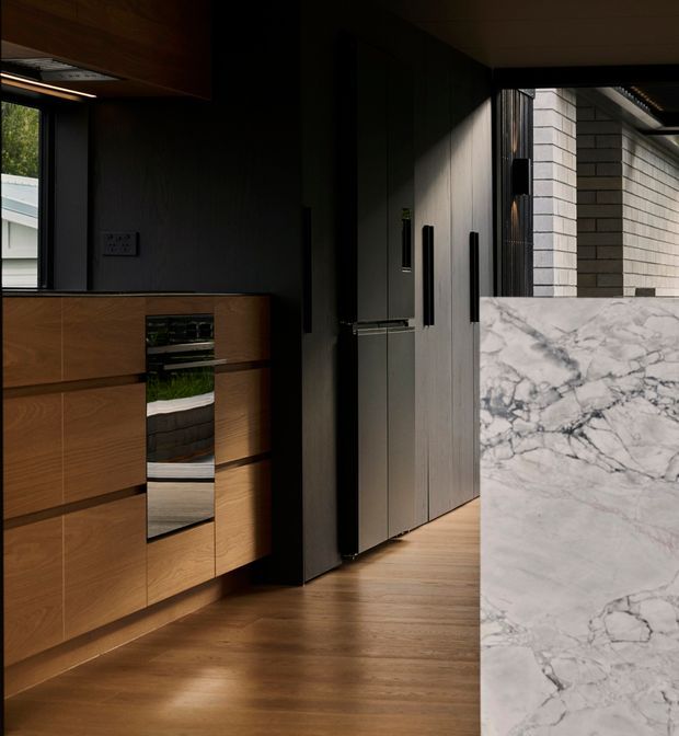 A dark, refined contemporary kitchen interior viewed toward a bank of tall integrated cabinetry. Warm timber cabinetry and timber flooring contrast with charcoal vertical panels, black hardware, and a large pale veined marble island or benchtop in the foreground. A window and open doorway provide limited natural light, with a glimpse of an exterior brick wall beyond.