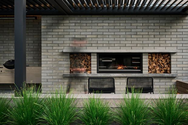 A contemporary outdoor entertaining area centered on a built-in grey brick fireplace with a lit fire, flanked by neatly stacked firewood niches. Two black woven lounge chairs sit on the paved terrace beneath a dark slatted overhead structure, with ornamental grasses in the foreground and a partially visible outdoor kitchen or barbecue at left.