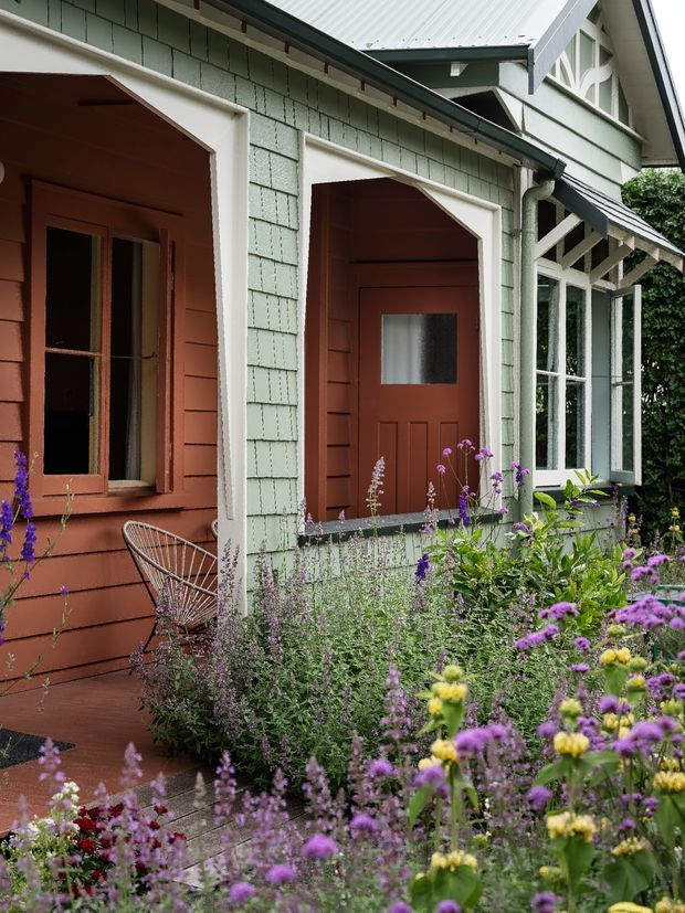 A close exterior view of a characterful cottage-style facade with pale green shingle cladding, terracotta-red timber wall sections, white trim, covered entry openings, timber doors, and a glazed veranda or sunroom. A lush cottage garden with abundant purple and yellow flowers fills the foreground.