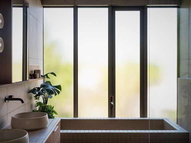 A serene modern bathroom featuring a deep built-in tiled soaking tub positioned beside large frosted floor-to-ceiling windows. The room includes a floating timber-toned vanity with round vessel basins, dark wall-mounted tapware, a frameless glass shower partition, integrated mirrored cabinetry, and abundant indoor greenery. The palette is composed of warm grey and beige tiles, dark metal frames, and natural wood tones, with soft diffused daylight creating a calm atmosphere.