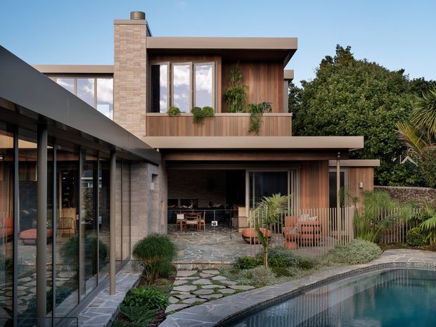 Exterior view of a contemporary two-storey residence arranged around a landscaped pool courtyard. The house features flat roof planes, extensive dark-framed glazing, warm vertical timber cladding, pale masonry walls, a covered outdoor dining or living area, stone paving, and lush planting.