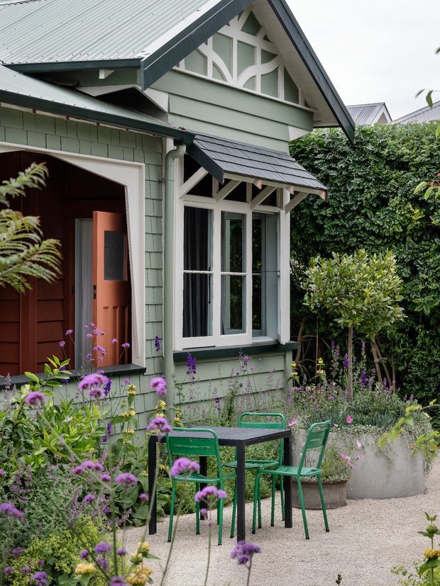 A close exterior view of a characterful gabled cottage or bungalow facade in a lush garden. The pale sage-green shingle-clad walls feature decorative white timber fretwork in the gable, a steep corrugated metal roof, a small dark slate-like awning over tall windows, and a warm timber entry door. A compact green metal outdoor dining set sits on a pale gravel courtyard surrounded by abundant flowering planting and hedges.