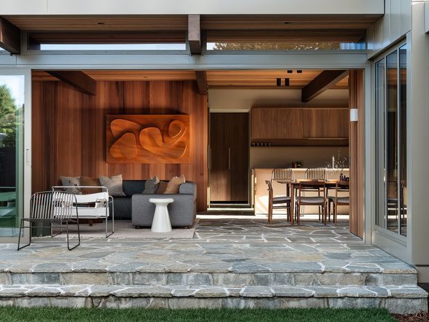 A sophisticated indoor-outdoor living area viewed from a stone terrace, with large open glazed doors connecting a wood-lined living room and dining/kitchen space. The living room features a grey sectional sofa, metal-framed lounge chair, sculptural wall art, and warm timber wall panelling. Beyond it is a dining table with chairs and built-in timber cabinetry. Broad stone steps lead down to a lawn, while clerestory glazing and exposed timber ceiling elements bring in natural light.