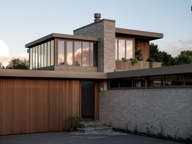 Exterior architectural photograph of a contemporary multi-level residence at dusk. The facade combines grey brick masonry, warm vertical timber cladding, dark metal roof planes and window frames, and extensive glazed areas. A projecting enclosed upper-level room, chimney, planted balcony or terrace, recessed entry, and low horizontal garage or ancillary wing are visible.